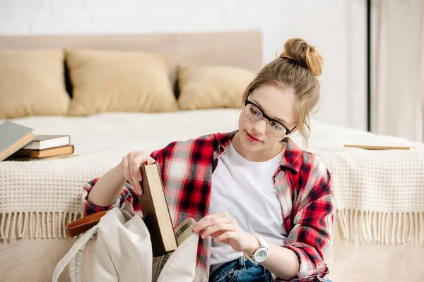 Teenager kid in glasses and checkered shirt putting book in backpack — Stock Photo
