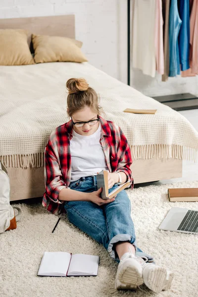 Adolescent concentré dans des lunettes et chemise à carreaux livre de lecture près du lit — Photo de stock