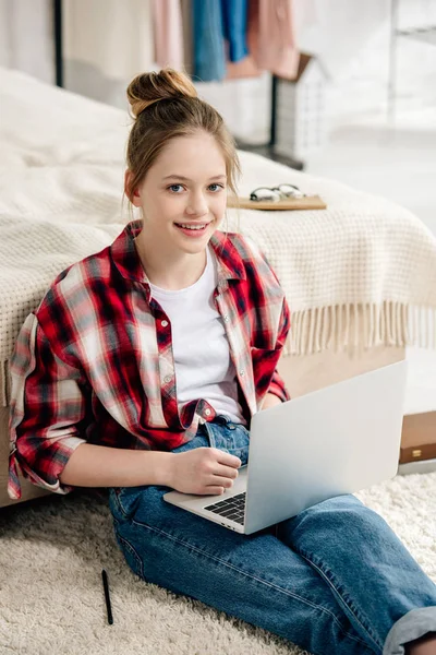 Smiling teenager in jeans sitting on carpet and using laptop — Stock Photo