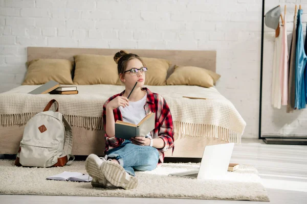 Adolescente con portátil y libros sentado en la alfombra y haciendo la tarea - foto de stock
