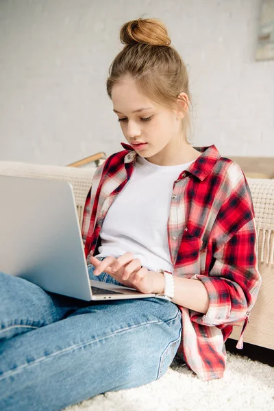 Teenage kid in checkered shirt using laptop while sitting on carpet — Stock Photo