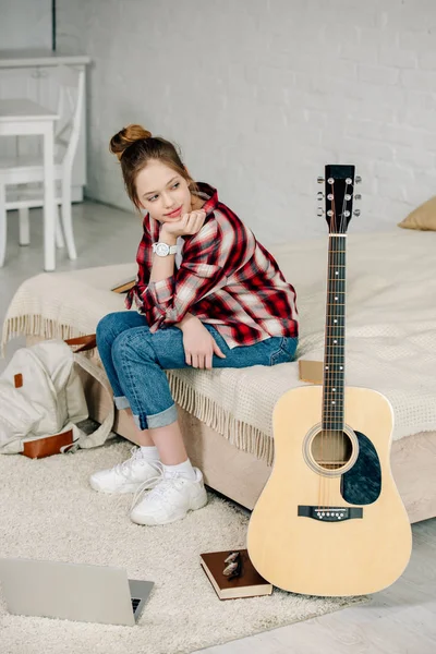Веселий підліток сидить на ліжку і дивиться на акустичну гітару — стокове фото