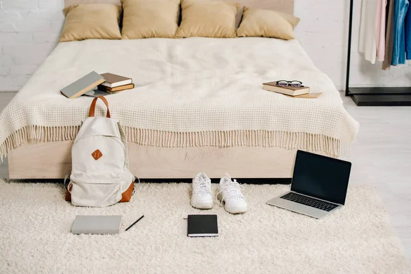 Zaino e laptop con schermo bianco sul tappeto vicino al letto — Foto stock