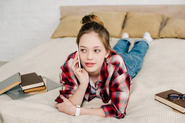 Teenager in checkered shirt lying on bed and talking on smartphone — Stock Photo