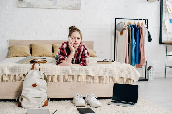 Teenager in checkered shirt lying on bed and talking on smartphone — Stock Photo