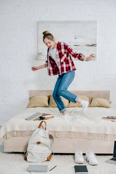 Visão completa do garoto adolescente sorridente em jeans pulando na cama em casa — Fotografia de Stock