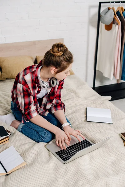 Teenager tippt im Kopfhörer auf Laptop-Tastatur, während er im Bett sitzt — Stockfoto
