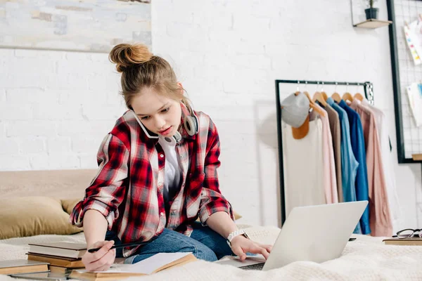 Adolescent inquiet assis sur le lit avec des livres et un ordinateur portable et parlant sur smartphone — Photo de stock