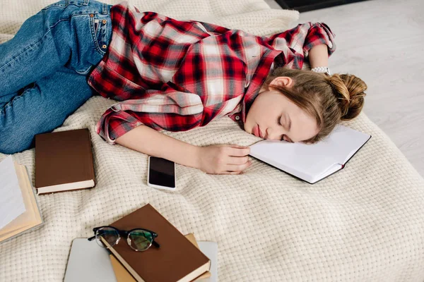 Підліток у картатій сорочці спить на ліжку з книгами та смартфонами — стокове фото