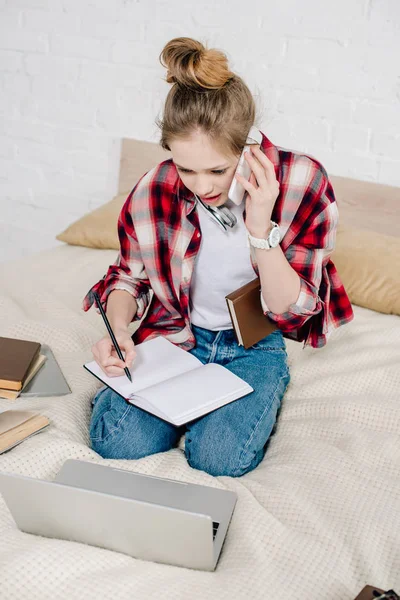 Worried teenager sitting on bed with books and laptop and talking on smartphone — Stock Photo