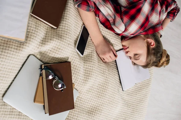 Підліток у картатій сорочці спить на ліжку з книгами та смартфонами — стокове фото