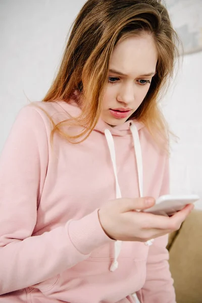 Niño adolescente sorprendido en sudadera con capucha rosa usando el teléfono inteligente en casa - foto de stock