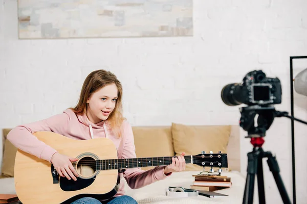 Blogger adolescente sentado en la cama y tocando la guitarra acústica frente a la cámara - foto de stock
