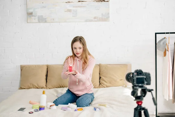 Blogger adolescente sentado en la cama y mostrando productos de belleza a la cámara - foto de stock