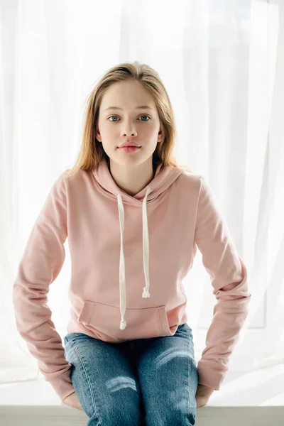 Front view of teenage kid in pink hoodie sitting on window sill and looking at camera — Stock Photo