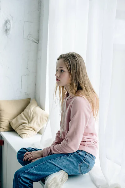 Pensive teenage kid in jeans sitting on window sill and looking away — Stock Photo