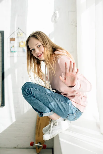 Sonriente adolescente en zapatillas de deporte sentado en el alféizar de la ventana y la mano agitada - foto de stock