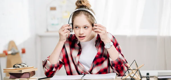 Panoramic shot of joyful teenage kid in checkered shirt listening music in headphones — Stock Photo
