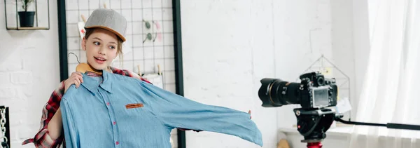Панорамный снимок веселого подростка в кепке, показывающего рубашку на видеокамеру — стоковое фото