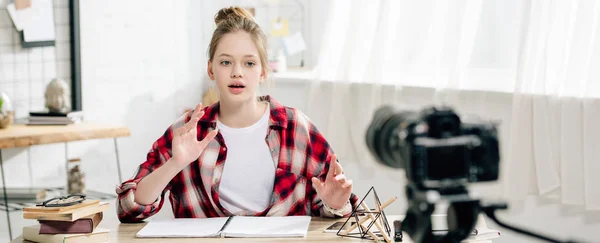 Panoramic shot of teenage blogger in checkered shirt sitting at table in front of video camera — Stock Photo