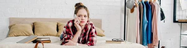 Panoramic shot of upset teenager in checkered shirt lying on bed and talking on smartphone — Stock Photo
