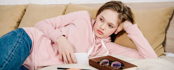 Panoramic shot of tired teenager with book and smartphone lying on bed — Stock Photo