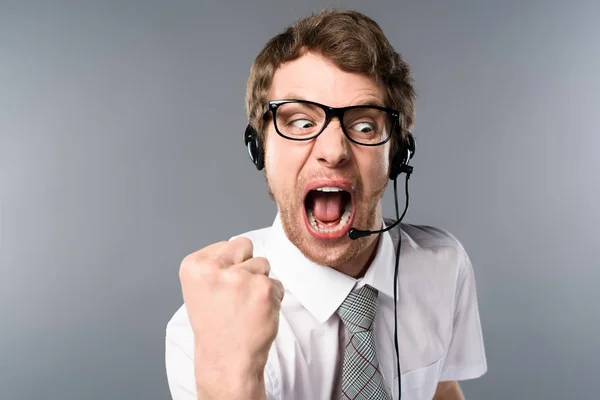 Operador de centro de llamadas enojado en auriculares y gafas gritando y mostrando el puño sobre fondo gris - foto de stock