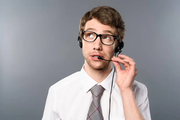 Operador de call center sonhador em fone de ouvido e óculos olhando para longe no fundo cinza — Fotografia de Stock