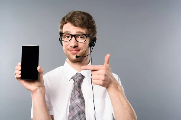 Operador del centro de llamadas positivo en gafas y auriculares que apuntan con el dedo al teléfono inteligente con pantalla en blanco - foto de stock