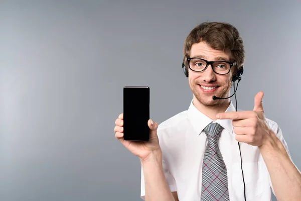 Lächelnder Callcenter-Betreiber mit Brille und Headset, der mit dem Finger auf Smartphone mit leerem Bildschirm zeigt — Stockfoto