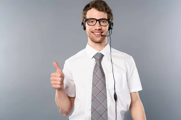 Smiling call center operator in glasses and headset showing thumb up on grey background — Stock Photo