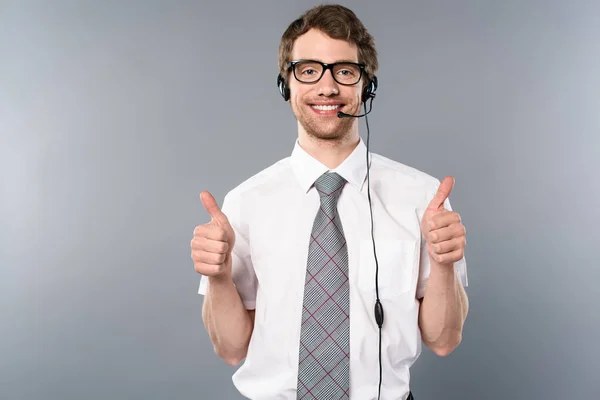 Smiling call center operator in glasses and headset showing thumbs up on grey background — Stock Photo