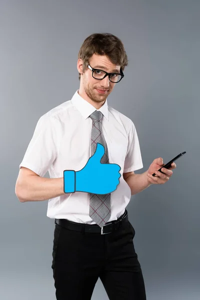 Businessman in glasses with paper cut thumb up using smartphone on grey background — Stock Photo