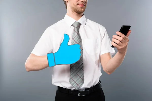 Vista recortada de hombre de negocios en gafas con el pulgar cortado de papel hacia arriba utilizando el teléfono inteligente sobre fondo gris - foto de stock