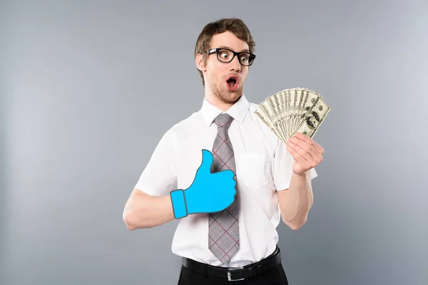 Sorprendido hombre de negocios en gafas con el pulgar cortado de papel hacia arriba la celebración de billetes de dólar sobre fondo gris — Stock Photo