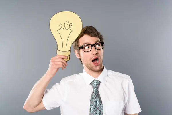 Hombre de negocios de ensueño con gafas que sostienen una bombilla de papel amarillo sobre fondo gris - foto de stock