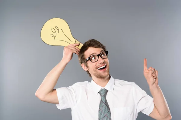 Happy businessman in glasses holding yellow paper cut light bulb and showing idea gesture on grey background — Stock Photo