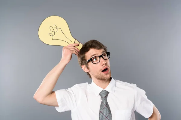 Dreamy businessman in glasses holding yellow paper cut light bulb and looking away on grey background — Stock Photo