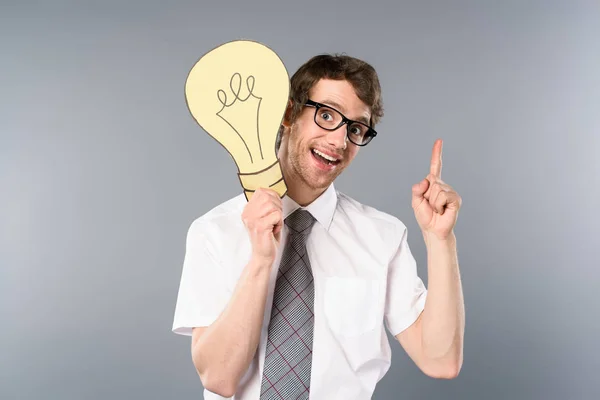 Улыбающийся бизнесмен в очках, держащий желтую бумагу, режет лампочку и показывает жест идеи на сером фоне — стоковое фото