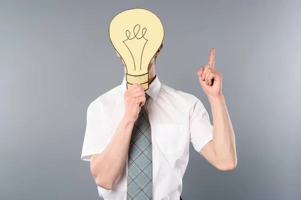 Businessman holding paper cut light bulb in front of face and showing idea gesture on grey background — Stock Photo