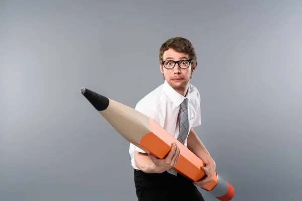 Tense businessman in glasses holding huge and heavy decorative pencil on grey background — Stock Photo