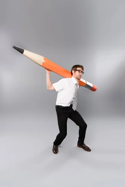 Funny tense businessman in glasses holding huge and heavy decorative pencil on grey background — Stock Photo