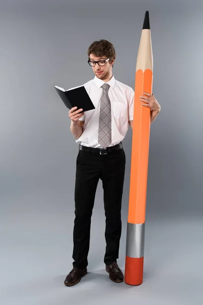 Concentrated businessman in glasses holding huge decorative pencil and reading book on grey background — Stock Photo