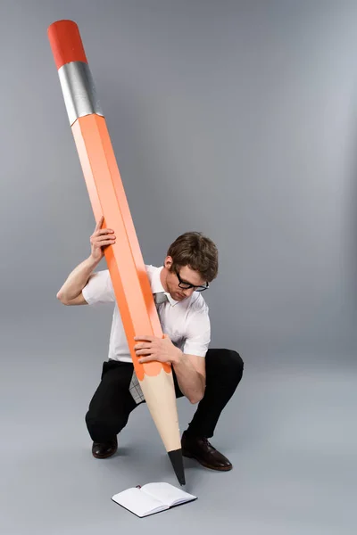 Geschäftsmann in Brille mit riesigem Zierstift und Notizbuch auf grauem Hintergrund — Stockfoto