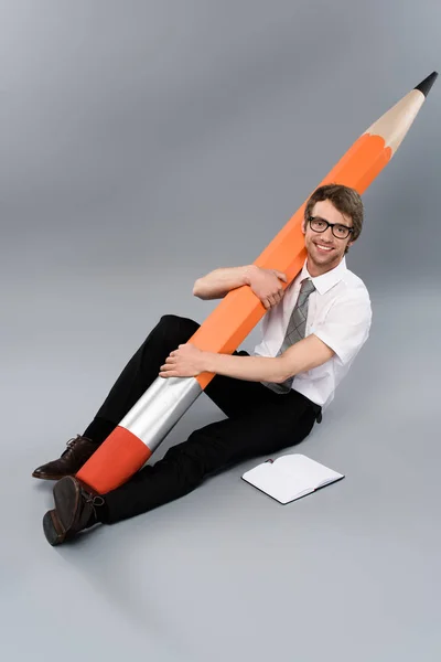 Happy businessman in glasses sitting on floor and holding huge decorative pencil on grey background — Stock Photo