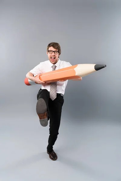 Drôle busines homme dans des lunettes tenant énorme crayon décoratif, criant et marchant sur fond gris — Photo de stock