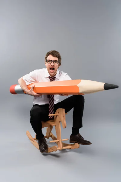 Funny businessman in glasses holding huge decorative pencil, screaming and riding wooden rocking horse on grey background — Stock Photo