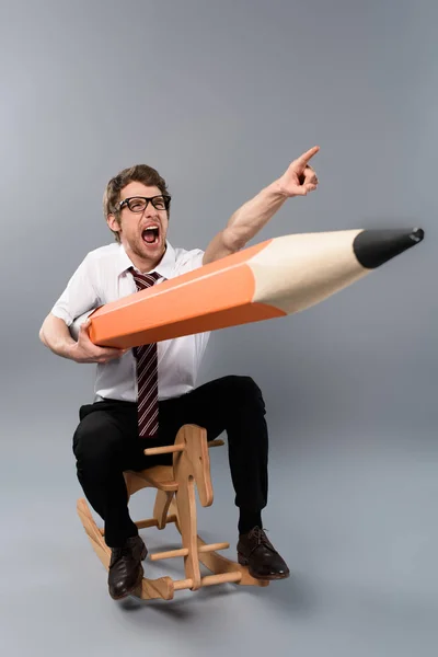 Funny businessman in glasses holding huge decorative pencil,pointing with finger, screaming and riding wooden rocking horse on grey background — Stock Photo