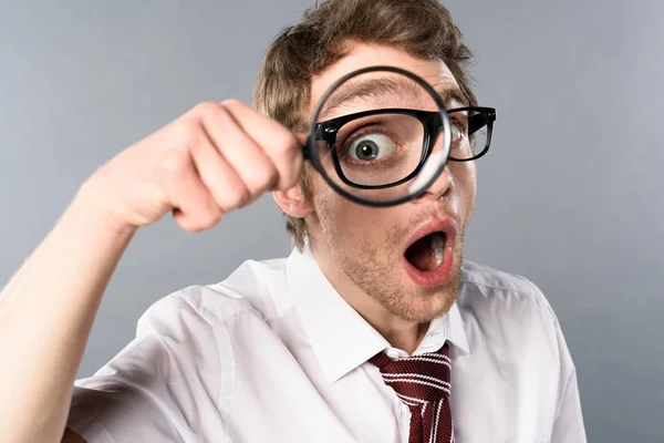 Empresário chocado em óculos com expressão facial engraçada olhando em lupa no fundo cinza — Fotografia de Stock