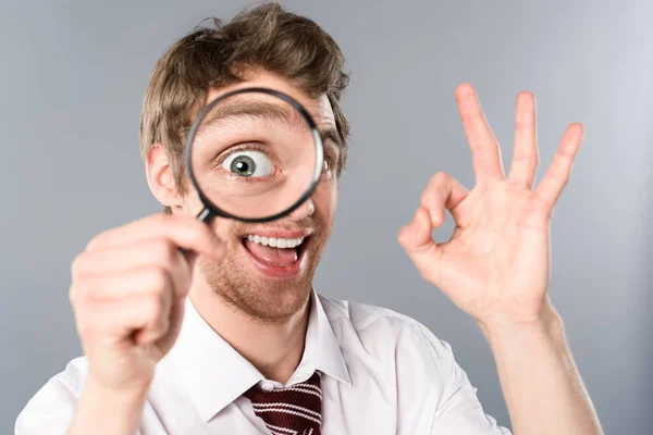 Homme d'affaires positif avec expression drôle de visage regardant dans la loupe et montrant ok signe sur fond gris — Photo de stock